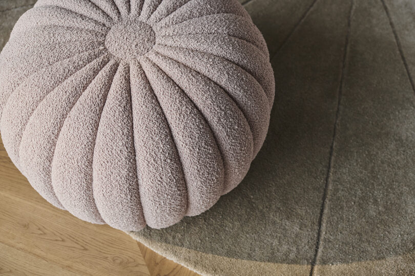 A textured, round ottoman with a ribbed design in soft pink rests on a tan rug with curved lines, set on a wooden floor.