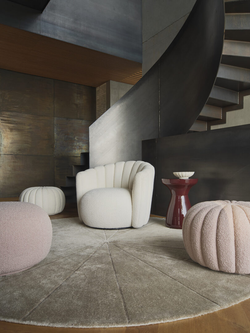 Modern interior with a white armchair, two round poufs, and a red side table on a circular rug. A curved staircase and textured walls complete the background.