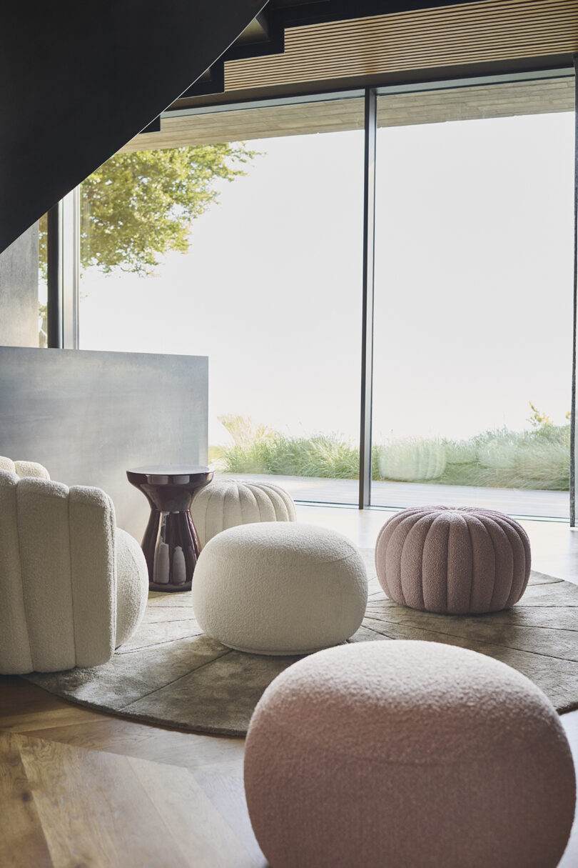 Modern living room with round poufs in white and pink tones on a circular rug. A small, black side table is nearby. Large windows offer a view of greenery outside.