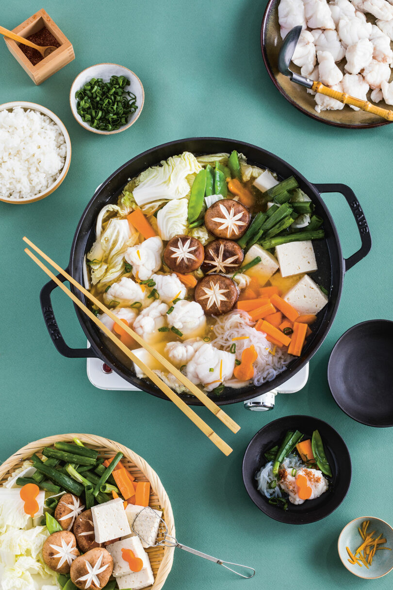 A pot of hot pot soup with vegetables, tofu, mushrooms, and seafood, surrounded by bowls of ingredients and garnishes, on a green table.