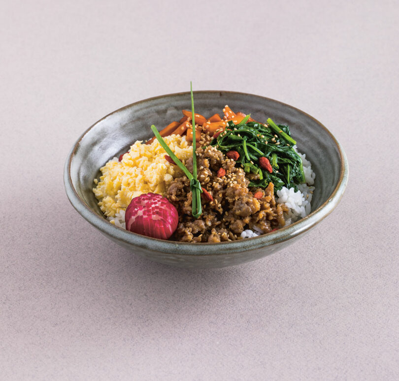 A bowl inspired by Sachiyo Harada, featuring rice topped with scrambled eggs, minced meat, cooked greens, diced carrots, a small red pickled item, and garnished with sesame seeds and a chive.