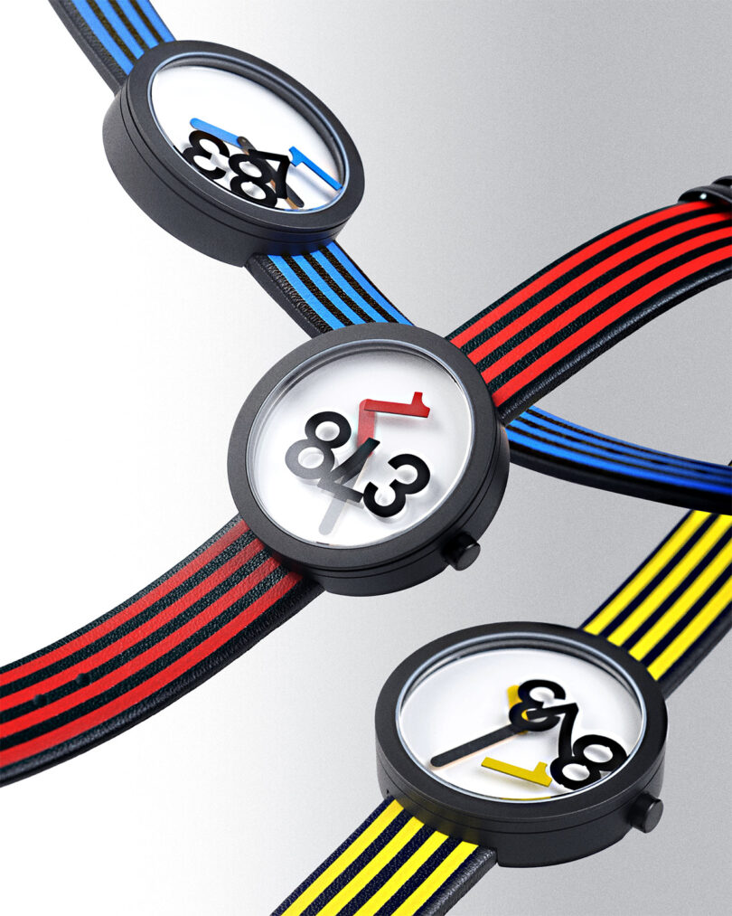 Three watches with black cases and striped bands in red, blue, and yellow. The dials display a large, tilted number design and minimalistic hour and minute hands.