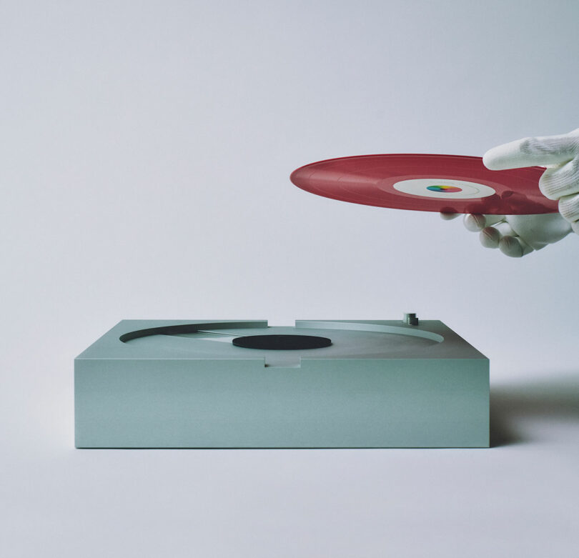 A gloved hand, reminiscent of the precision associated with PP-1 components, holds a red vinyl record above a minimalist turntable against a plain background.