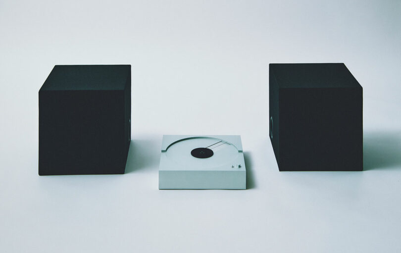 A sleek PP-1 white record player takes center stage, flanked by two imposing black speakers on a minimalist backdrop.