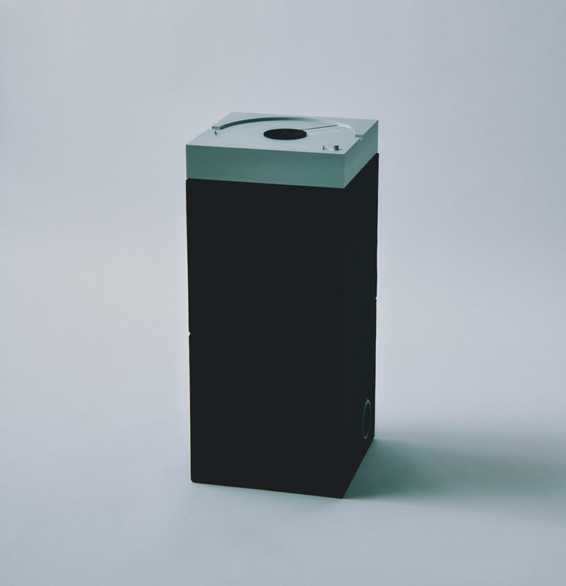 The PP-1 speaker stands tall and sleek with its rectangular black body and a striking silver top, set elegantly against a plain backdrop.