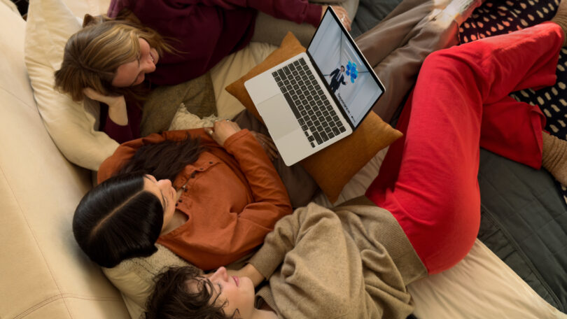 Three people lying on a bed, watching something on a laptop placed on a pillow.