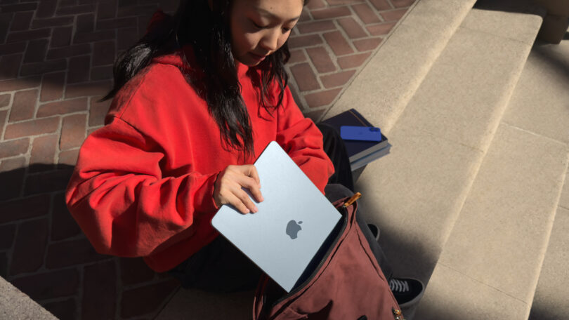 Person in a red sweater sitting on steps, placing a closed laptop with an Apple logo into a maroon bag.