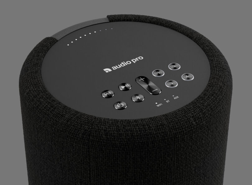 Black cylindrical speaker with a fabric cover, featuring control buttons and an "audio pro" logo on top against a plain gray background.