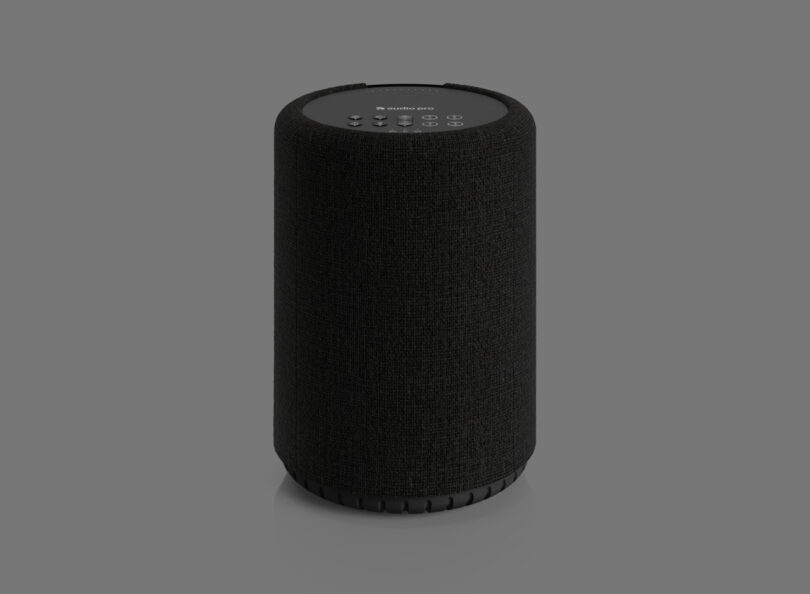 Black cylindrical smart speaker with a textured surface, featuring buttons on the top panel, set against a plain gray background.