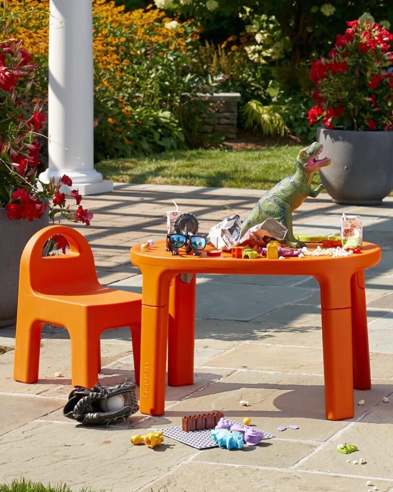 Orange kids' table and chair outdoors with toy dinosaurs, snacks, and scattered items on and around the table, surrounded by vibrant red flowers