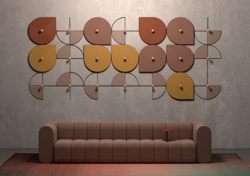 A modern brown sofa sits on a rug below a wall installation featuring geometric shapes in brown, orange, and yellow tones. A small red side table is attached to the sofa