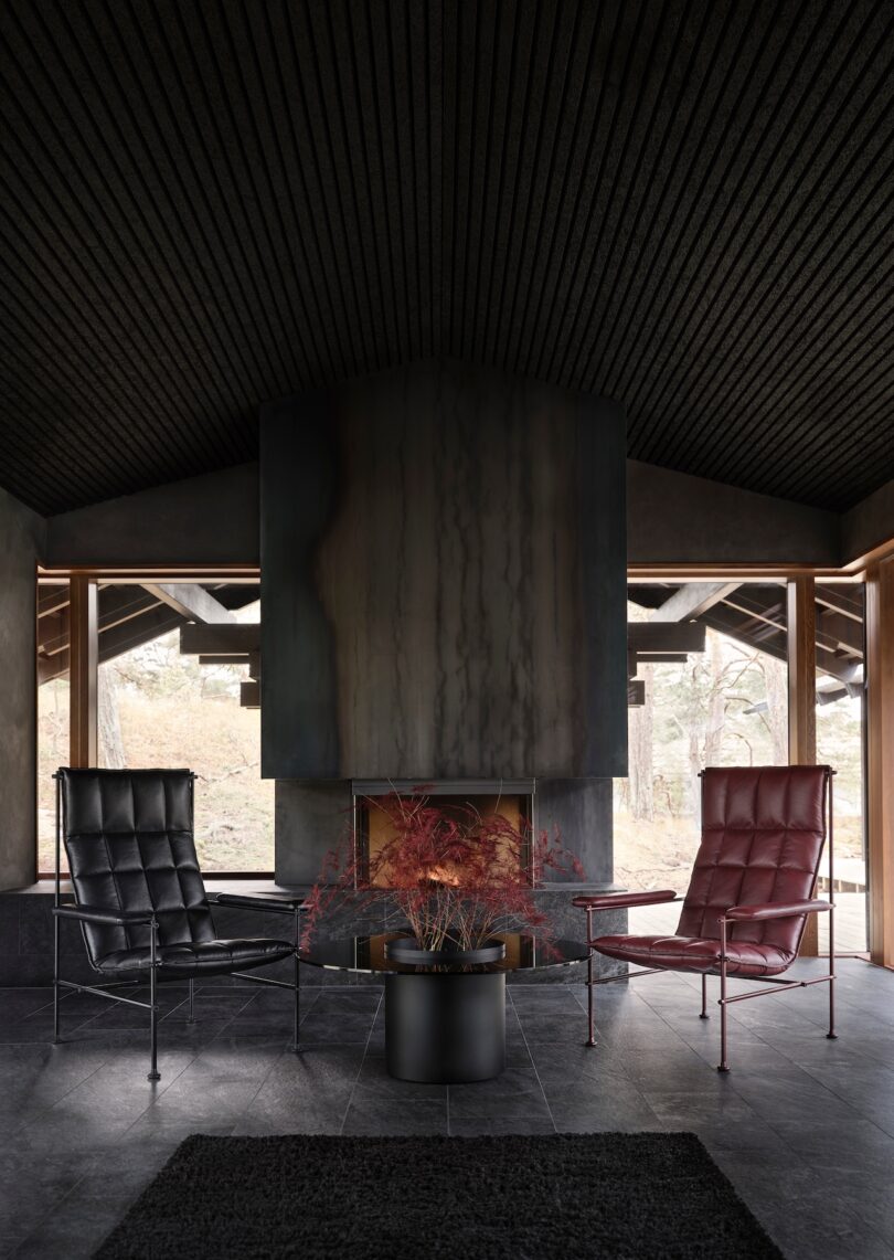Modern living room with a dark color scheme, featuring a black chair, a red chair, a central fireplace, and a pot with red foliage on a black-tiled floor. Large windows provide natural light