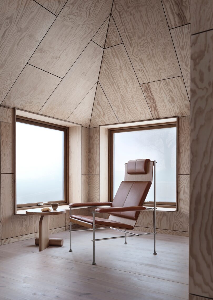 Minimalist room with wooden walls and floor, featuring a brown leather chair with a headrest and metal frame. A small wooden side table holds a bowl, situated between two large windows