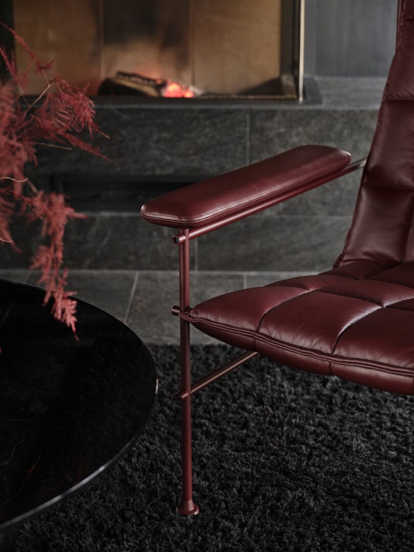A dark brown leather armchair with a metal frame sits on a black rug near a round black table. A fireplace with a glow is visible in the background