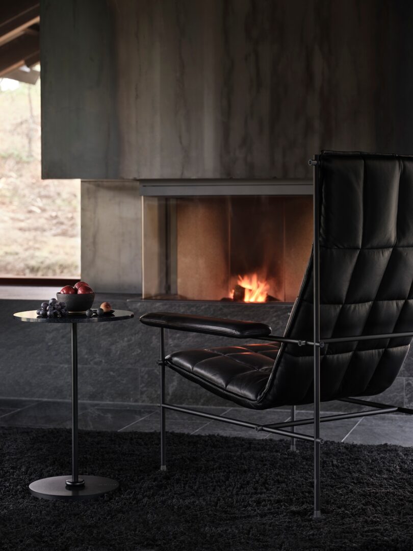 A black leather chair faces a lit fireplace. A small round table beside the chair holds fruit. The room is dimly lit with a view of the outdoors through a window