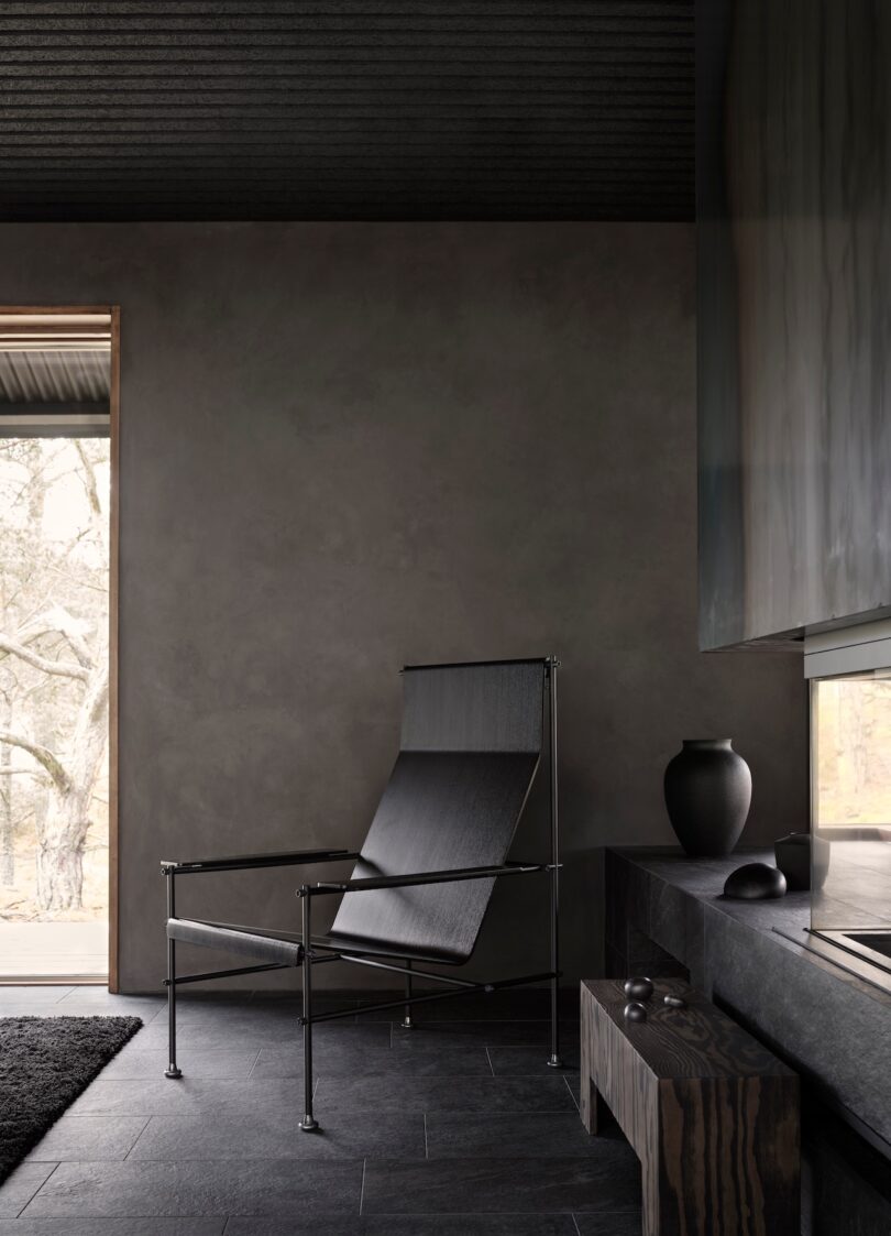 Minimalist interior with a black chair, textured dark walls, and a large window showing a tree outside. Dark furniture includes a low table with a vase and a small round object