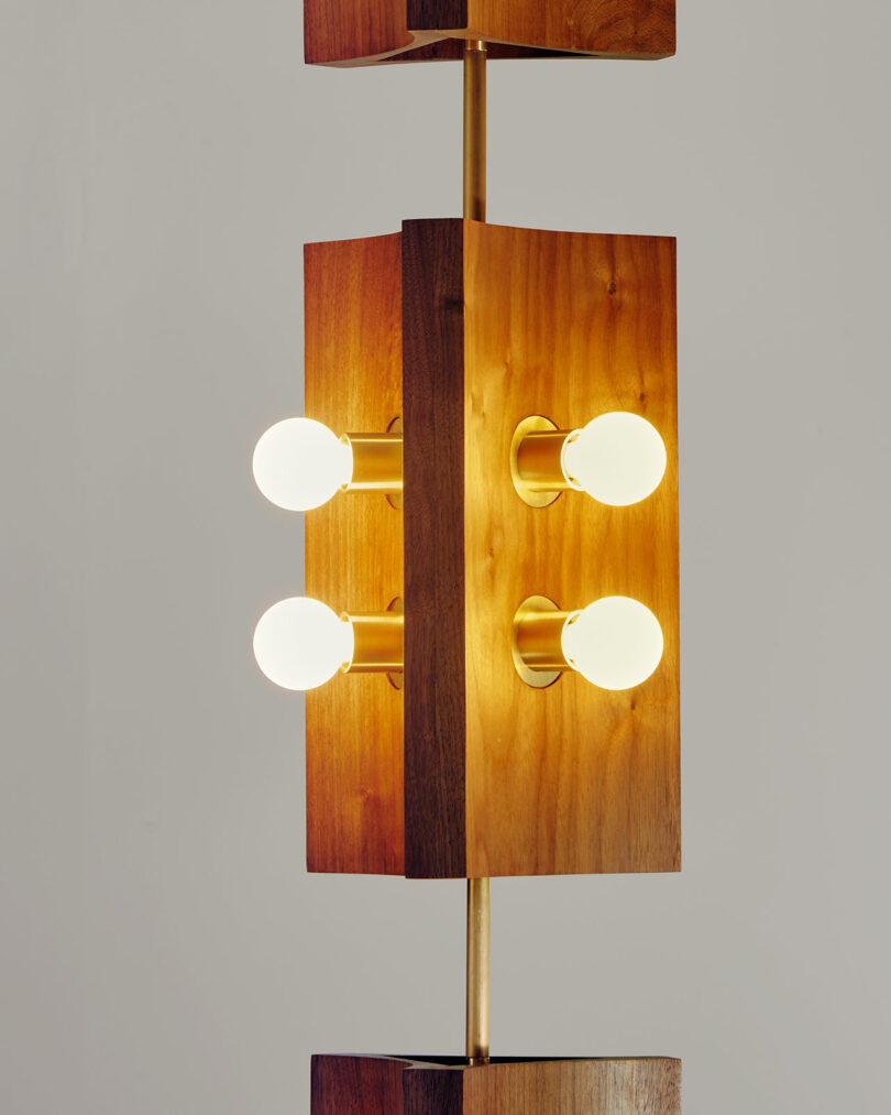 A tall wooden lamp from Blue Green Works features four illuminated bulbs on a central vertical rod, set against a plain background.