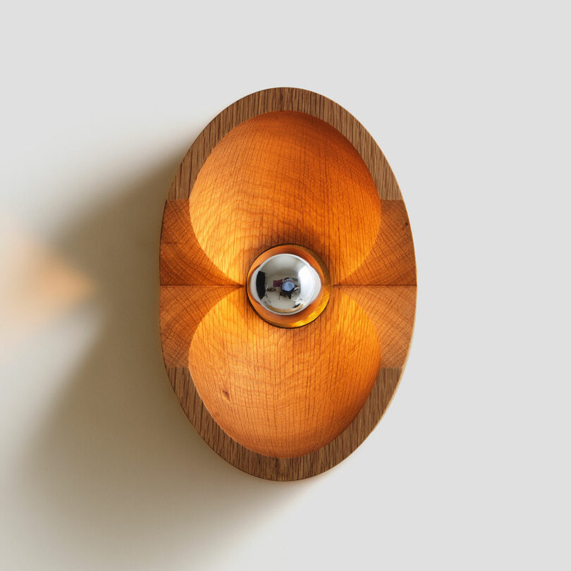 A round wooden wall light fixture from Blue Green Works emits a warm glow, with a chrome central bulb casting geometric shadows on the wall.