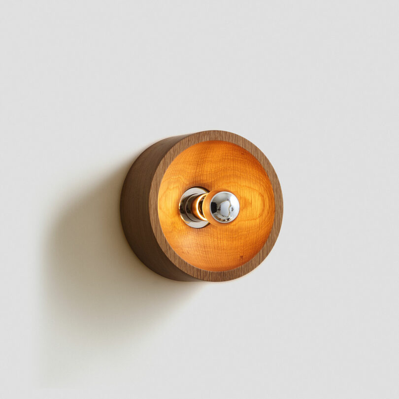 A round wooden wall lamp with a spherical chrome bulb, mounted on a white wall. Crafted by Blue Green Works, the lamp emits a warm, orange glow.