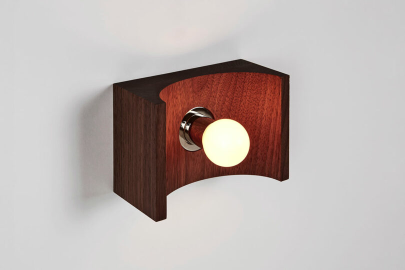 The minimalist wooden wall sconce by Blue Green Works features a single exposed bulb, elegantly mounted on a plain wall.