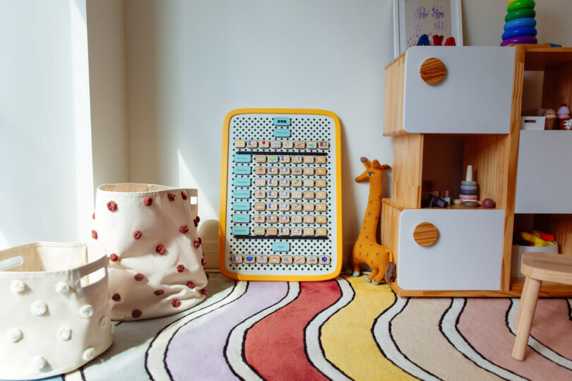 The colorful children's room, reminiscent of a Design Milk Take 5 feature, showcases a toy abacus, giraffe, and polka dot baskets on a vibrant wavy rug. A sleek white and wooden storage unit with round handles sits by the window, bathing in bright light