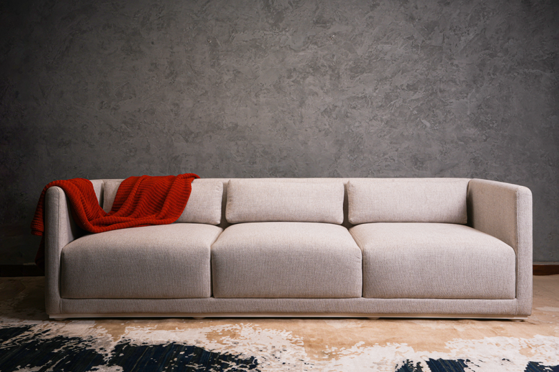 A beige, modern sofa with clean lines, adorned with a red throw blanket, set against a textured gray wall on a rug-covered floor