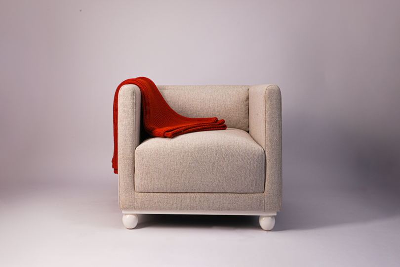 A beige armchair with rounded arms and white legs, draped with a folded red blanket on the left side, is set against a plain, light-colored background