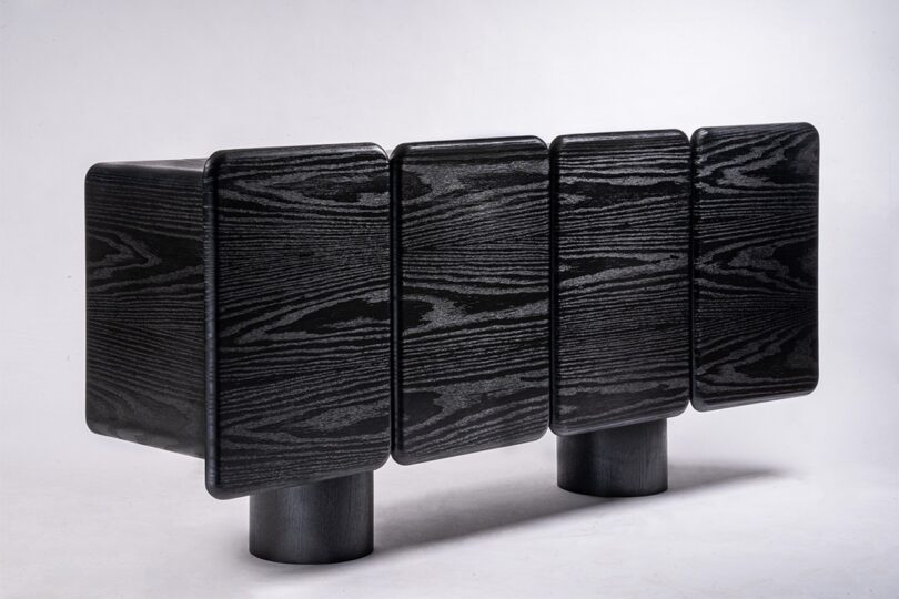 A black, rectangular wooden cabinet with four textured panels and two cylindrical legs on a white background