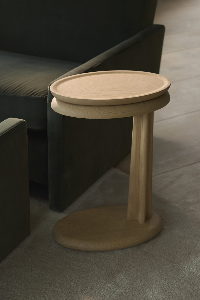 A wooden side table with a round top stands beside a dark green upholstered sofa on a carpeted floor
