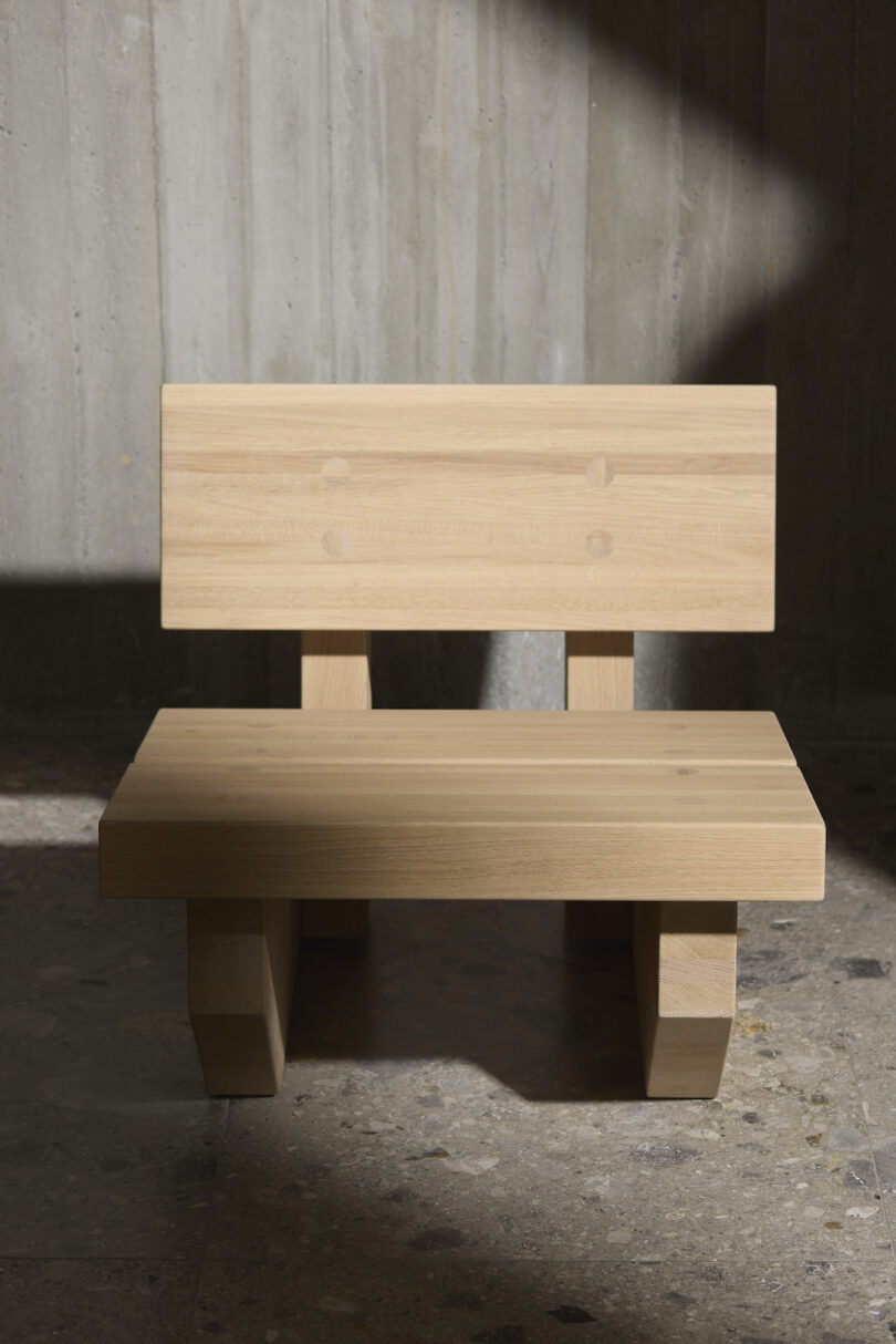 Minimalist wooden chair with a straight backrest and flat seat, placed on a stone floor against a concrete wall, partially in shadow