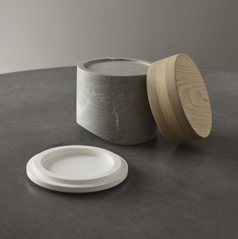 A gray cylindrical container with a separate wooden lid stands on a dark surface, next to a round white plate