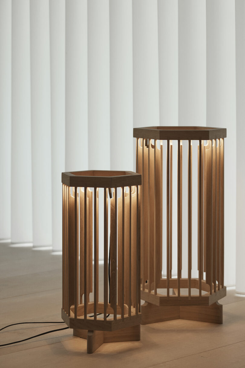 Two wooden floor lamps with vertical slats stand on a light wood floor, illuminated against a backdrop of white vertical blinds