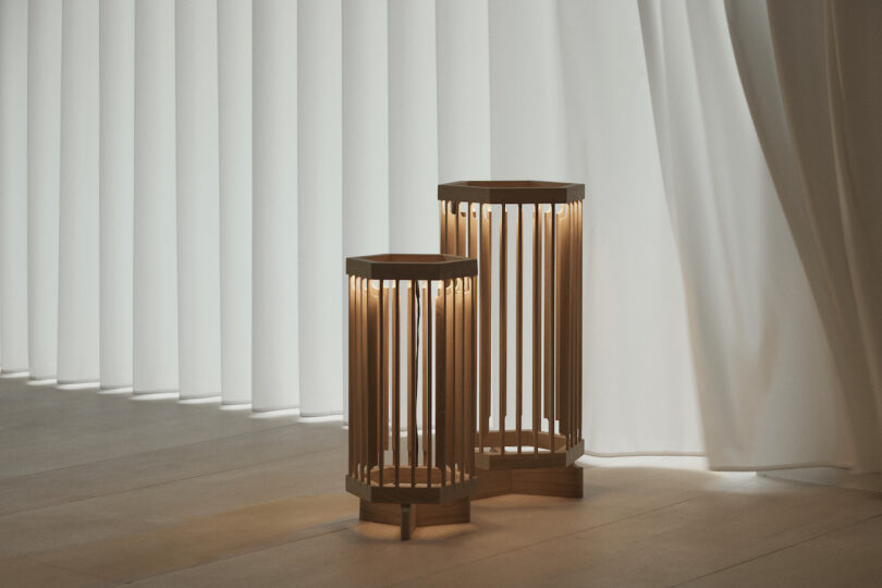 Two wooden, lantern-style lights on a wooden floor beside curtains, casting warm light