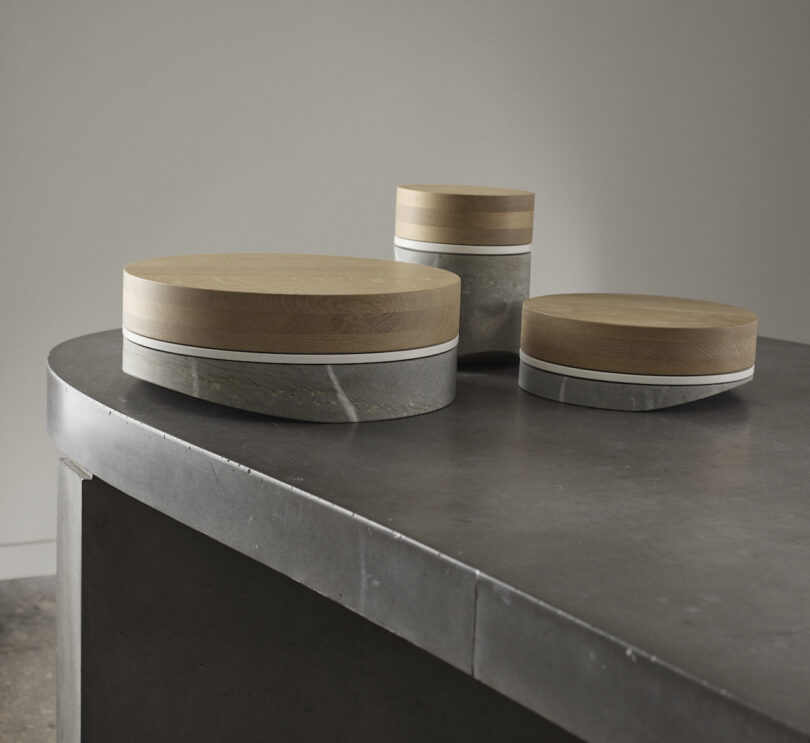 Three round wooden and metal containers with lids are placed on a curved, dark stone surface