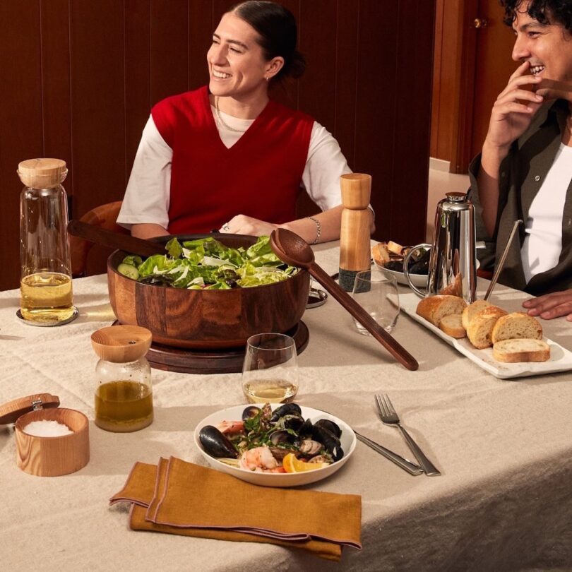 Two people seated at a dining table with a large salad bowl, bread, glasses, and a dish with mussels