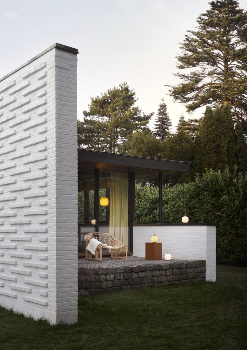 A modern house with a white brick exterior features a glass wall, wicker chairs on the patio, and radiant Louis Poulsen portable lamps. Surrounded by trees and greenery, it offers an inviting blend of nature and contemporary design