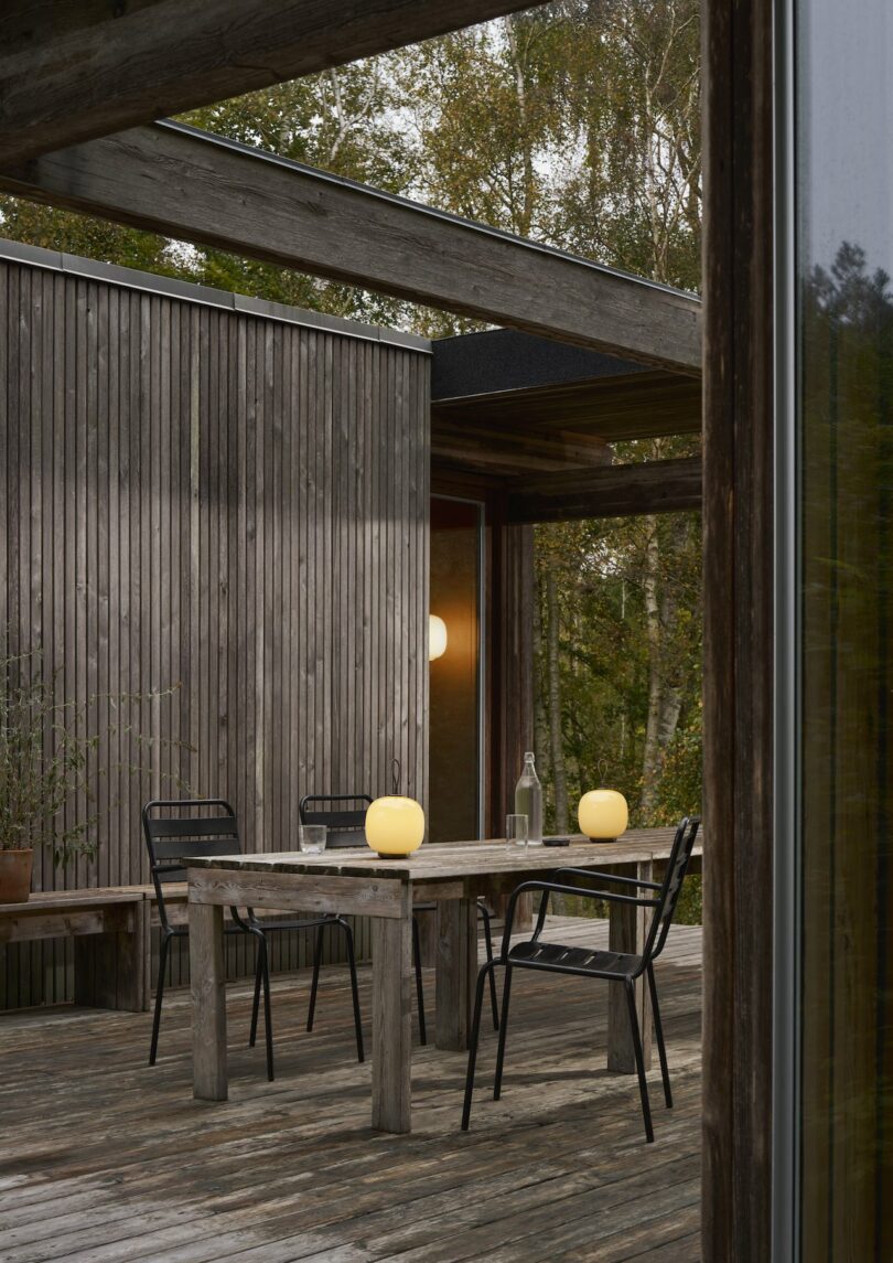 An inviting outdoor wooden patio features a table and three chairs illuminated by two Louis Poulsen portable lamps. A glass bottle completes the serene setup, all nestled against rustic wooden walls with trees in the background