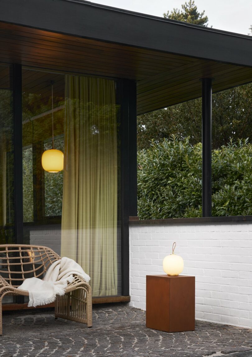 A rattan bench with a cozy blanket sits next to a wooden cube topped with a sleek Louis Poulsen portable lamp. Green curtains drape over large windows, while a brick wall frames the patio, offering glimpses of trees in the background