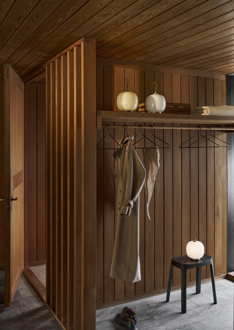 Wood-paneled room with an open door, a coat and hat hanging on a rack, a bench illuminated by a Louis Poulsen Portable Lamp casting warm light, and neatly folded towels positioned above