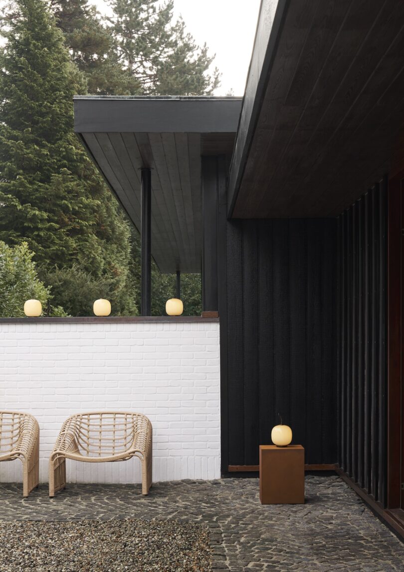 A modern house exterior boasts a black and white color scheme. On the stone patio, two wicker chairs invite relaxation, while a Louis Poulsen Portable Lamp adds a touch of elegance alongside round lights on the wall. Trees create a serene backdrop