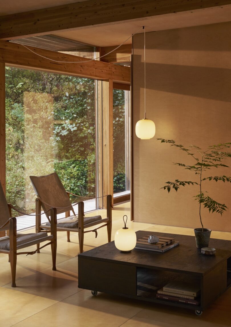 Minimalist room with two wooden chairs, a coffee table featuring a Louis Poulsen portable lamp, books, and a plant. Large windows reveal greenery outside, while ceiling and table lamps provide warm lighting