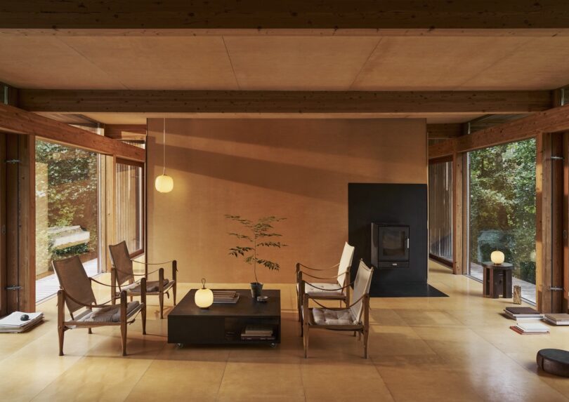 A minimalist living room showcases wooden beams, large windows, two chairs, a coffee table, and a wood stove. The decor is complemented by a small plant, spherical pendant lights, and a Louis Poulsen Portable Lamp for added elegance