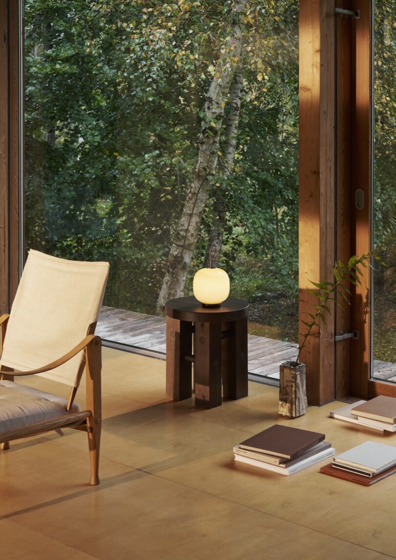 A minimalistic room features a wooden chair and a small round table holding a Louis Poulsen portable lamp that casts a warm glow. Several stacked books sit nearby, with large windows showcasing a lush green forest outside