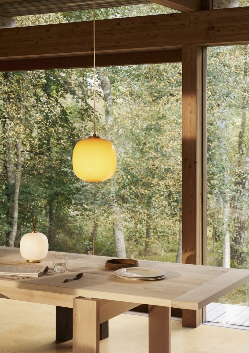 Dining table with two place settings under a Louis Poulsen Portable Lamp, next to large windows showcasing a stunning forest view