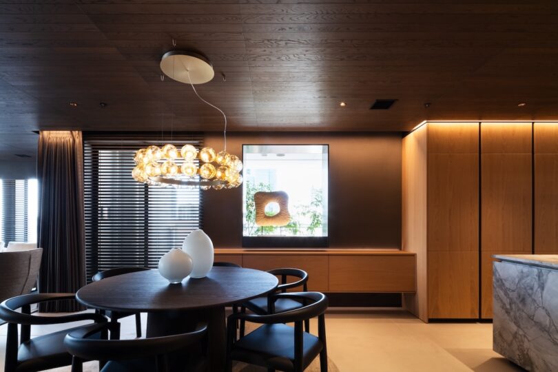 Modern dining area with a round table, black chairs, and a chandelier. Wooden cabinets and blinds, with a decorative piece on the back wall, complement the contemporary interior design.