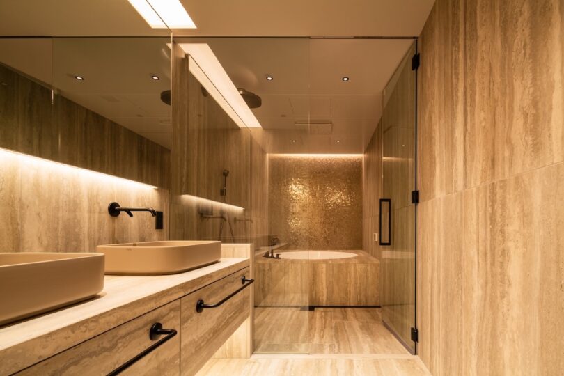 Modern bathroom with beige marble tiles, dual sinks, black fixtures, a large mirror, and a built-in bathtub with ambient lighting.