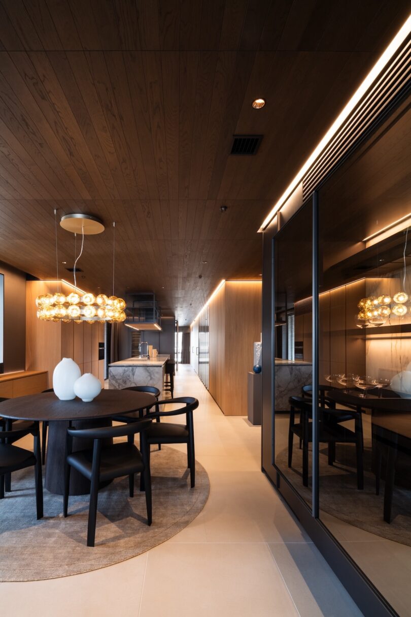 Modern dining area with a round table, dark chairs, and a decorative light fixture. Warm lighting highlights the wood ceiling and wall panels.