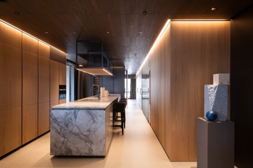 Modern kitchen with a sleek marble island, wooden cabinetry, and ambient lighting. A pedestal with sculptures is on the right.