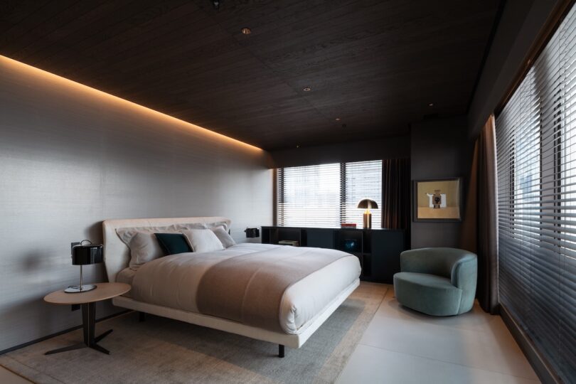 Modern bedroom with a large bed, side table with lamps, cushioned chair, and blinds covering the windows. Walls and ceiling are dark-toned, with soft lighting above the bed.