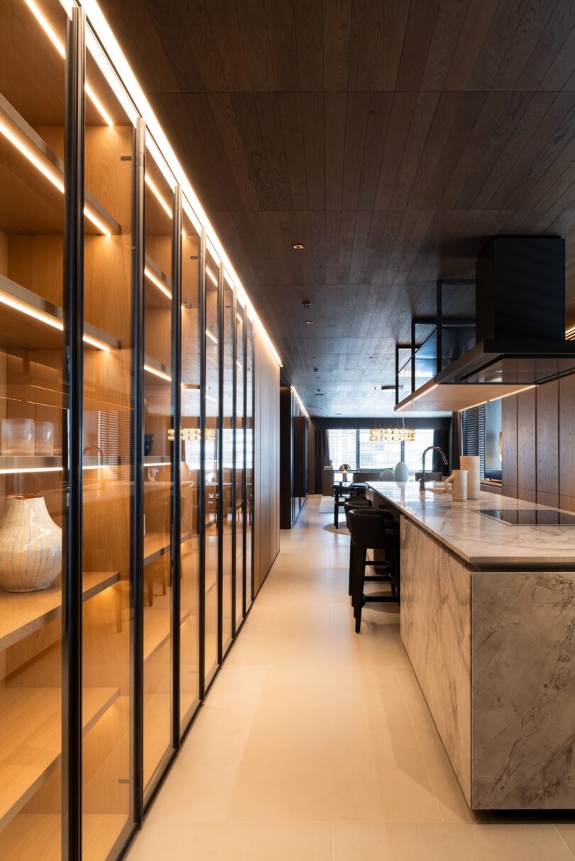 Modern kitchen with marble island, wooden cabinets with glass doors, and recessed lighting. Neutral tones and sleek design create a minimalist aesthetic.